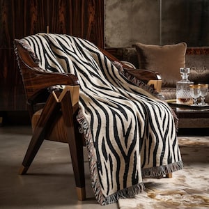 Pode incluir: Uma manta tecida com um padrão de riscas de zebra em preto e creme com extremidades em franjas, drapeada sobre uma poltrona de madeira escura com detalhes em metal dourado. A manta exibe um design de estampado animal de alto contraste.