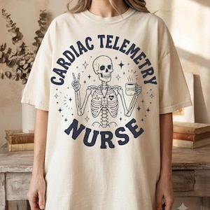 May include: An off-white short-sleeve t-shirt featuring a dark blue graphic of a skeleton making a peace sign and holding a coffee mug. The text "CARDIAC TELEMETRY NURSE" is printed in a circular layout around the skeleton, accented by small stars.