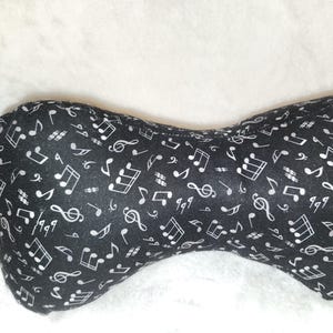 Pode incluir: Uma almofada preta em forma de osso coberta por um padrão de notas musicais brancas, claves de sol e outros símbolos musicais. A almofada tem um pequeno laço de tecido. Projetada para suporte de pescoço.