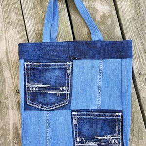 Könnte beinhalten: Eine Denim-Tragetasche aus verschiedenen Blautönen. Die Tasche hat zwei Vordertaschen mit weißen Nähten und einen dunkelblauen Denim-Oberteil. Die Griffe sind aus dem gleichen Denim-Material.