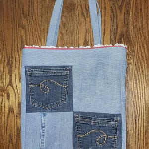 May include: A blue denim tote bag with a white lace trim and red stitching. The bag is made from recycled denim and has four pockets.