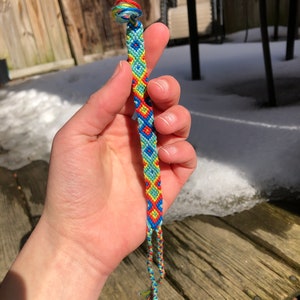 May include: A vibrant friendship bracelet featuring a diamond pattern in blue, red, yellow, and green. The handmade bracelet has a knotted thread design, a loop at one end, and braided ties at the other.