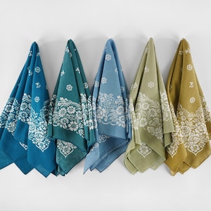 May include: Five colorful bandanas hanging against a white wall. Each bandana features a floral design in white against a different color: teal, dark teal, light blue, olive green, and mustard yellow. The bandanas are folded into triangles.