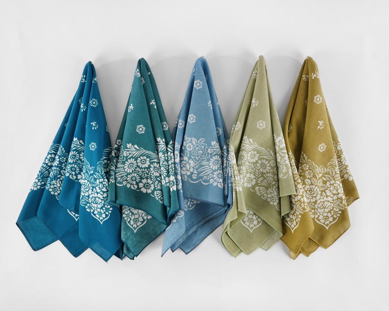 May include: Five colorful bandanas hanging against a white wall. Each bandana features a floral design in white against a different color: teal, dark teal, light blue, olive green, and mustard yellow. The bandanas are folded into triangles.
