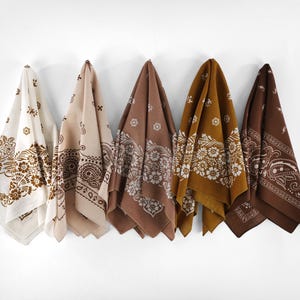 May include: Five bandanas in varying shades of brown and cream hang against a white wall. Each bandana features a different floral or paisley pattern. The bandanas are made of a soft fabric.