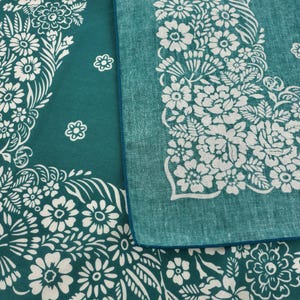 May include: Two teal bandanas with white floral patterns. One bandana is folded, revealing a contrasting blue edge. The floral design includes various flower shapes and leaf details, creating a decorative border.
