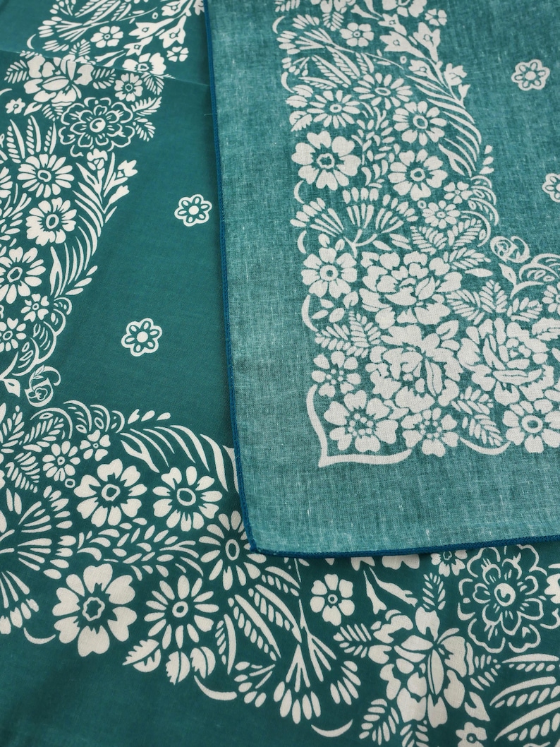 May include: Two teal bandanas with white floral patterns. One bandana is folded, revealing a contrasting blue edge. The floral design includes various flower shapes and leaf details, creating a decorative border.