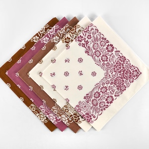 May include: A collection of five square fabric napkins in shades of brown, mauve, and cream. Each napkin features floral patterns, with some showcasing all-over designs and others displaying a decorative border. The napkins are arranged in a fan shape.