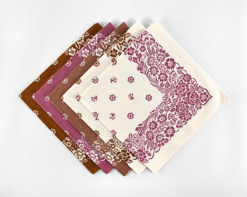 May include: A collection of five square fabric napkins in shades of brown, mauve, and cream. Each napkin features floral patterns, with some showcasing all-over designs and others displaying a decorative border. The napkins are arranged in a fan shape.
