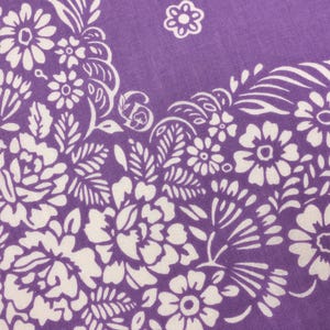 May include: Close-up of a purple bandana with a white floral pattern. The design features detailed white flowers, leaves, and swirling accents. The fabric has a soft, textured appearance, and the overall aesthetic is vintage and decorative.