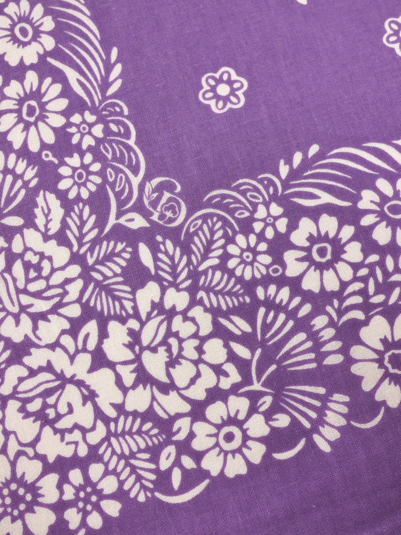 May include: Close-up of a purple bandana with a white floral pattern. The design features detailed white flowers, leaves, and swirling accents. The fabric has a soft, textured appearance, and the overall aesthetic is vintage and decorative.