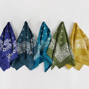 May include: Five bandanas in shades of blue, teal, green, and yellow hang against a white background. Each bandana features a white floral and paisley design. The bandanas are made of a soft fabric.
