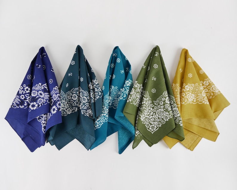 May include: Five bandanas in shades of blue, teal, green, and yellow hang against a white background. Each bandana features a white floral and paisley design. The bandanas are made of a soft fabric.
