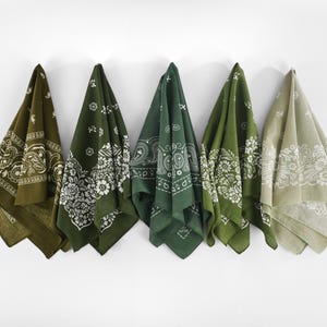 May include: Five green bandanas with white paisley patterns hanging against a white wall. The bandanas vary in shades of green, from olive to forest green, and are folded to display their designs.