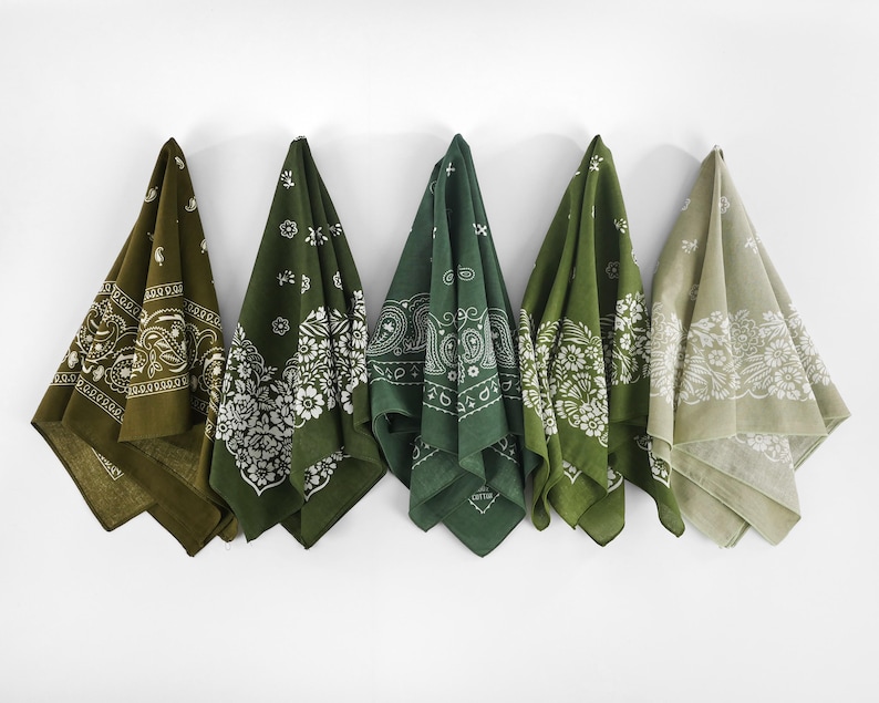 May include: Five green bandanas with white paisley patterns hanging against a white wall. The bandanas vary in shades of green, from olive to forest green, and are folded to display their designs.
