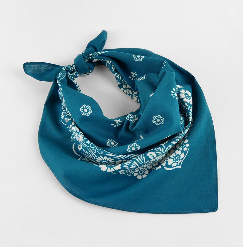 May include: Teal bandana with a white floral pattern. The square scarf is knotted at the top, showcasing the detailed design. The edges feature a repeating floral motif, creating a classic accessory.