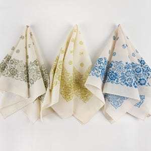 May include: Three off-white bandanas with floral patterns in olive green, yellow-green, and blue. Each bandana is folded and hanging against a white background. The floral designs are printed along the edges of the bandanas.