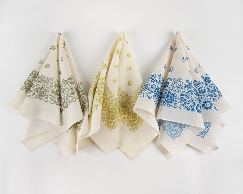 May include: Three off-white bandanas with floral patterns in olive green, yellow-green, and blue. Each bandana is folded and hanging against a white background. The floral designs are printed along the edges of the bandanas.