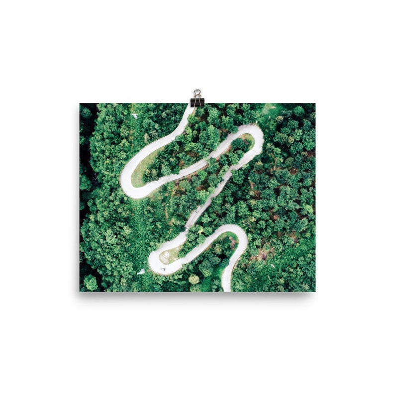 May include: An aerial view of a winding white road through a dense green forest. The road curves and twists through the trees, creating a unique and interesting pattern.