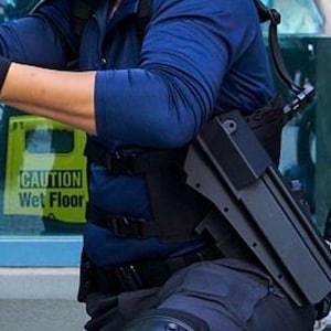 May include: A dark blue long-sleeved shirt worn with a black tactical vest and a black holster attached to the side. The outfit includes black gloves and dark trousers. A yellow sign with the black text CAUTION Wet Floor is visible in the background.