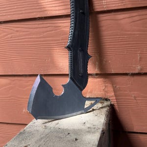 May include: A black, stylized axe with a curved handle and a sharp blade. The axe is resting on a concrete surface against a reddish-brown wooden wall. The handle has a textured grip.