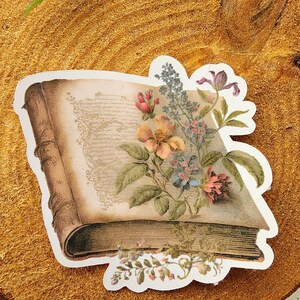 May include: A vintage book sticker with a floral design. The book is open and has a floral arrangement on top of it. The flowers are in shades of pink, purple, and yellow.