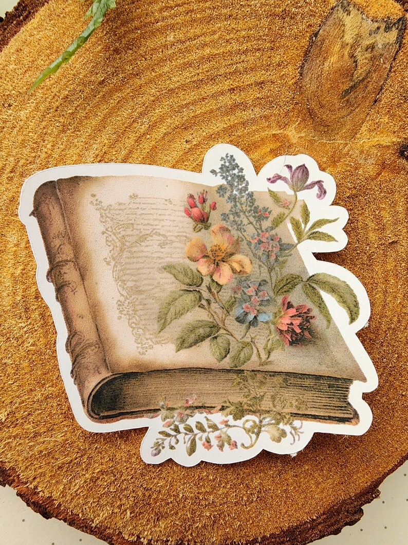 May include: A vintage book sticker with a floral design. The book is open and has a floral arrangement on top of it. The flowers are in shades of pink, purple, and yellow.