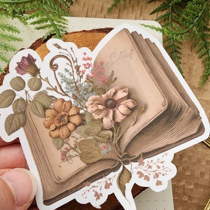 May include: A vintage book sticker with a floral design. The book is open and has a floral arrangement on top of it. The flowers are in shades of pink, brown, and white.
