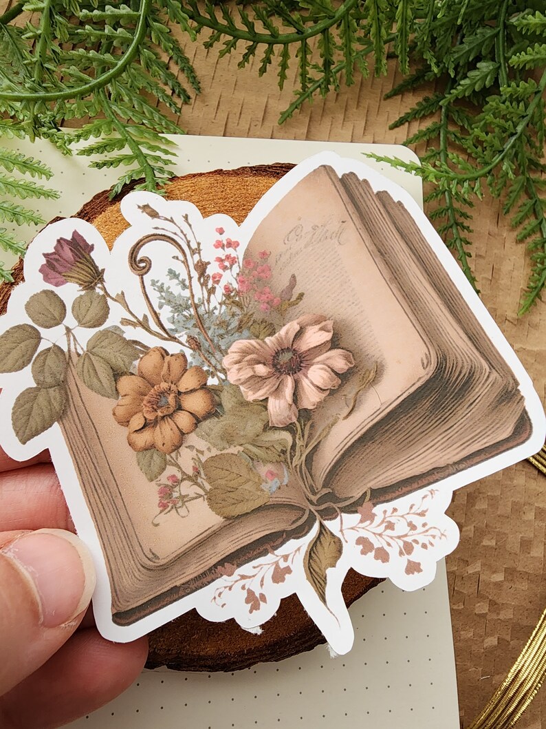 May include: A vintage book sticker with a floral design. The book is open and has a floral arrangement on top of it. The flowers are in shades of pink, brown, and white.