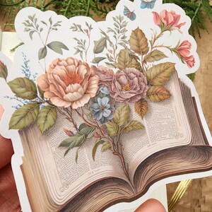 May include: A vintage style sticker featuring an open book with a bouquet of flowers, including roses, blue flowers, and green leaves. A blue butterfly is flying above the flowers. The book has text on the pages and a brown cover.