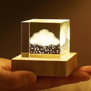 May include: A small, clear glass cube with a white cloud and raindrops inside, lit from within. The cube sits on a wooden base.