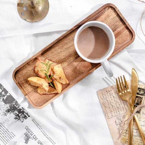 Puede incluir: Una bandeja de madera con una taza blanca llena de una bebida marrón, gajos de patata y cubiertos dorados. La bandeja es rectangular con esquinas redondeadas. Una ramita de romero se encuentra encima de los gajos de patata.