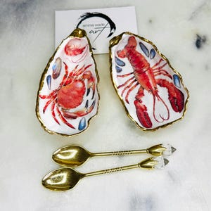 May include: Two decorative oyster shell dishes with gold rims, one featuring a red crab and the other a red lobster. Two gold spoons with crystal accents are also present. The image includes a white card with black text.