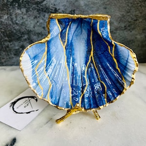 May include: A blue and gold seashell dish with a gold stand. The seashell is painted with a blue and white marbled design and accented with gold lines.