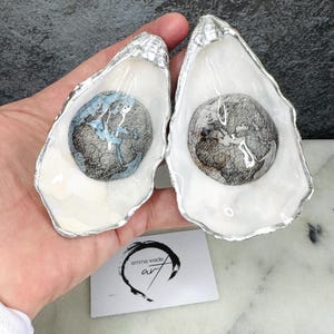 May include: Two oyster shells with silver edging, each holding a globe design. One globe is blue and white, the other is grey and black. A small card with "emma wade art" is visible.