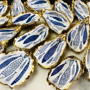 Luxury hand gilded Dorset oyster shell trinket dish featuring a refined blue fish motif, edged in liquid gold leaf and sealed in crystal clear resin. Sustainable coastal ring holder handmade by the artist in Surrey and presented in eco friendly branded gift packaging. Elegant beach house decor piece ideal for jewellery storage, wedding styling or sophisticated coastal interiors.