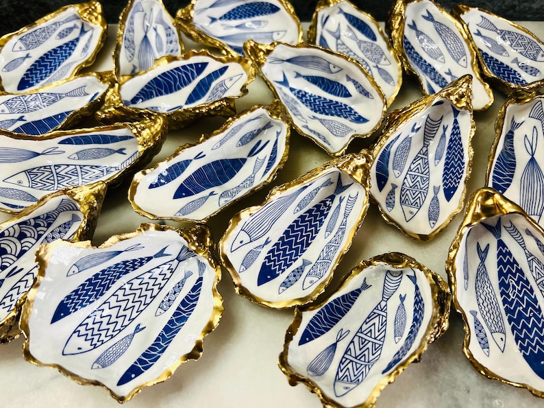 Luxury hand gilded Dorset oyster shell trinket dish featuring a refined blue fish motif, edged in liquid gold leaf and sealed in crystal clear resin. Sustainable coastal ring holder handmade by the artist in Surrey and presented in eco friendly branded gift packaging. Elegant beach house decor piece ideal for jewellery storage, wedding styling or sophisticated coastal interiors.