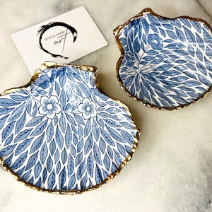 May include: Two blue and white ceramic clam shell dishes with gold trim. The shells have a floral pattern with blue leaves and white flowers.