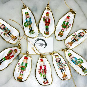 Peut inclure: Ornements de Noël avec des motifs de casse-noisettes. Chaque décoration est peinte sur une base blanche en forme de coquillage, avec une bordure dorée et un fil doré pour suspendre. Les ornements présentent des casse-noisettes aux couleurs festives.