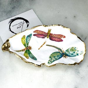 Handmade dragonfly oyster shell trinket dish with hand-gilded gold leaf edges and glossy resin finish, created from sustainably sourced Dorset shells. Unique coastal jewellery holder and ring dish, ideal as a vanity tray, trinket tray or decorative keepsake. Nature inspired home decor gift for birthdays, weddings, anniversaries, housewarmings and Mother’s Day. One of a kind artisan shell dish, beautifully presented in a gift box.