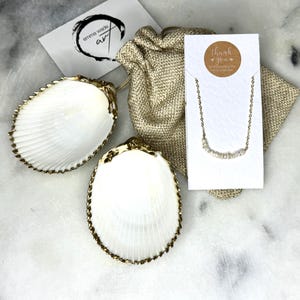 May include: A gold necklace with a row of small white pearls, displayed on a white card with the text "Thank you". The necklace is packaged in a small burlap bag.