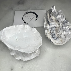 May include: Two white oyster shell dishes. One is a single shell, the other is a stack of three shells tied with a silver ribbon. The card in the background reads "emma wade art".