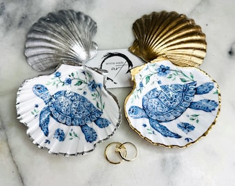 May include: Two decorative scallop shells featuring a blue and white sea turtle illustration with floral accents. One shell has a silver-painted edge, the other a gold-painted edge. A small white card with the text emma wade art and two gold rings.