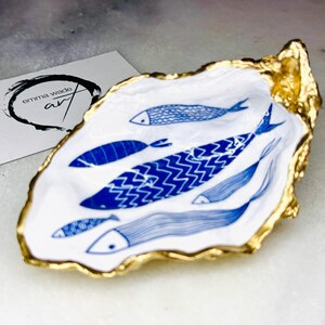 Luxury hand gilded Dorset oyster shell trinket dish featuring a refined blue fish motif, edged in liquid gold leaf and sealed in crystal clear resin. Sustainable coastal ring holder handmade by the artist in Surrey and presented in eco friendly branded gift packaging. Elegant beach house decor piece ideal for jewellery storage, wedding styling or sophisticated coastal interiors.