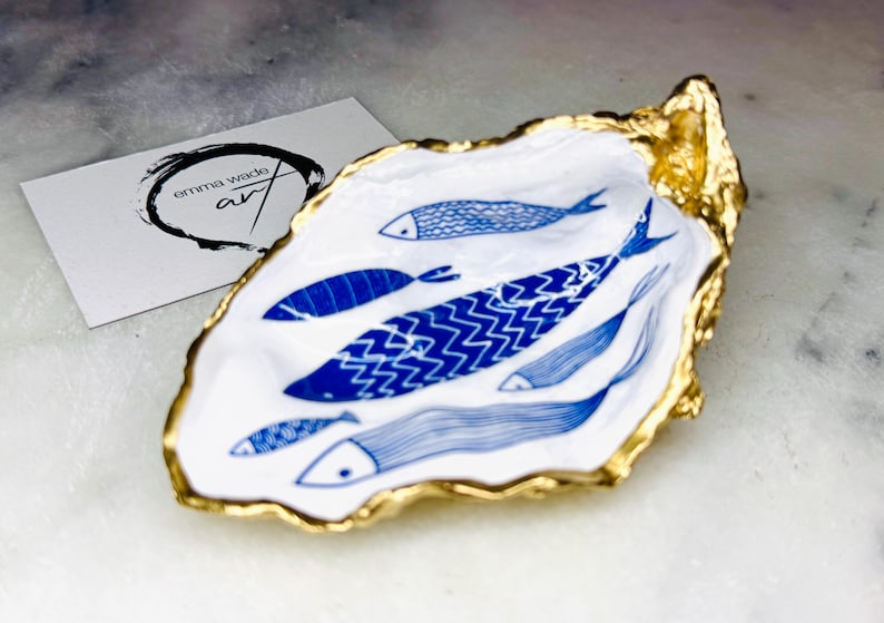 Luxury hand gilded Dorset oyster shell trinket dish featuring a refined blue fish motif, edged in liquid gold leaf and sealed in crystal clear resin. Sustainable coastal ring holder handmade by the artist in Surrey and presented in eco friendly branded gift packaging. Elegant beach house decor piece ideal for jewellery storage, wedding styling or sophisticated coastal interiors.