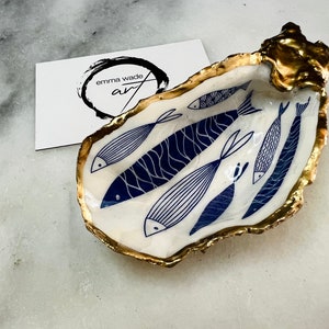 Hand-gilded Dorset oyster shell trinket dish featuring a blue fish motif, edged with liquid gold leaf and sealed in high gloss resin. Coastal ring holder and nautical home decor piece displayed with multiple shells. Sustainable oyster shell art handmade in Surrey, ideal as a jewellery dish, wedding favour, beach house accessory or ocean lover gift.