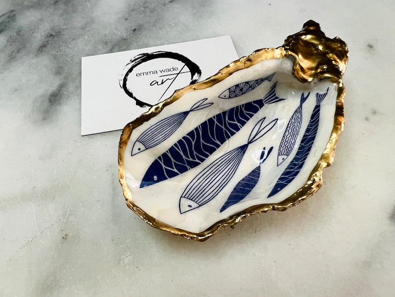 Hand-gilded Dorset oyster shell trinket dish featuring a blue fish motif, edged with liquid gold leaf and sealed in high gloss resin. Coastal ring holder and nautical home decor piece displayed with multiple shells. Sustainable oyster shell art handmade in Surrey, ideal as a jewellery dish, wedding favour, beach house accessory or ocean lover gift.