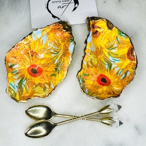 Può includere: Due piccoli piatti a forma di conchiglia di ostrica dorati con un motivo dipinto di girasoli. I piatti sono su una superficie di marmo bianco. Sono incluse due cucchiai d'oro con manici in cristallo trasparente.