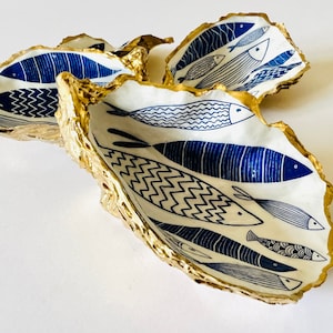 Hand-gilded Dorset oyster shell trinket dish featuring a blue fish motif, edged with liquid gold leaf and sealed in high gloss resin. Coastal ring holder and nautical home decor piece displayed with multiple shells. Sustainable oyster shell art handmade in Surrey, ideal as a jewellery dish, wedding favour, beach house accessory or ocean lover gift.