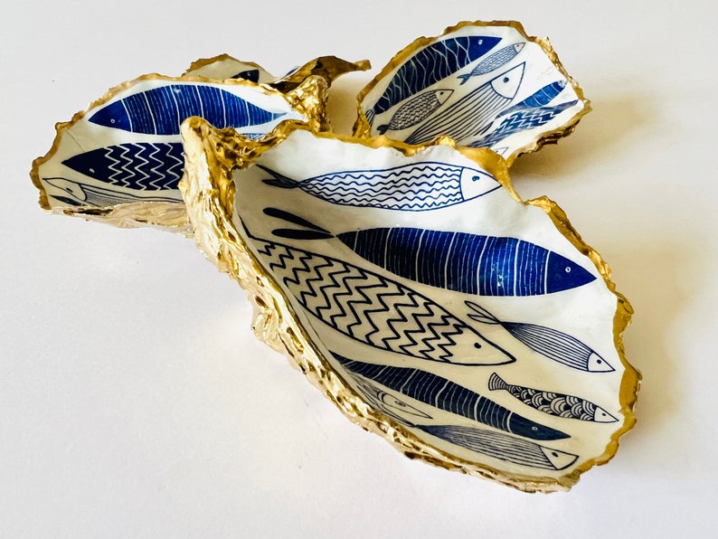 Hand-gilded Dorset oyster shell trinket dish featuring a blue fish motif, edged with liquid gold leaf and sealed in high gloss resin. Coastal ring holder and nautical home decor piece displayed with multiple shells. Sustainable oyster shell art handmade in Surrey, ideal as a jewellery dish, wedding favour, beach house accessory or ocean lover gift.